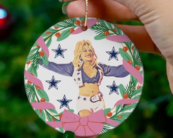 May include: A white ceramic Christmas ornament featuring a woman wearing a blue and white jersey with stars, a pink bow, and a green wreath with pink ribbons. The ornament is decorated with green leaves and red berries.