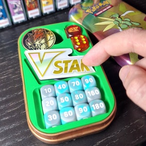 May include: A green and copper-coloured Pokémon V STAR dice and coin set. The set includes dice, a coin, and numbered dice. The background shows a collection of Pokémon game cases.