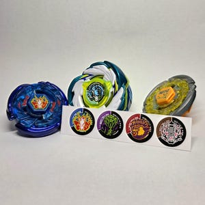 Pu&ograve; includere: Quattro trottole colorate con vari disegni e una striscia di adesivi abbinati. Le trottole presentano sfumature di blu, verde, giallo e arancione. Gli adesivi presentano illustrazioni dettagliate di creature e simboli mitici.