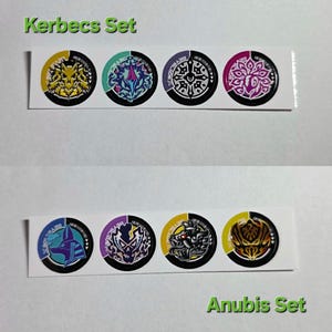 Pu&ograve; includere: Sono esposti due set di adesivi circolari. Il set superiore, etichettato "Kerbecs Set", presenta quattro disegni con accenti gialli, verde acqua, viola e rosa. Il set inferiore, "Anubis Set", mostra disegni con colori blu, viola, giallo e oro.