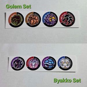 Pu&ograve; includere: Sono esposti due set di adesivi circolari. Il set superiore, etichettato "Golem Set", presenta quattro adesivi con vari disegni di mostri in arancione, marrone, viola e rosso. Il set inferiore, etichettato "Byakko Set", presenta anch'esso quattro adesivi con disegni di mostri in viola, rosso, blu e bianco.