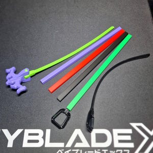 Peut inclure: Ensemble d'accessoires de jouets colorés, comprenant des cordons de tirage verts, violets, rouges, noirs et verts. Un lanceur violet est visible à gauche. Le texte "YBLADE X" et des caractères japonais sont imprimés sur la surface.