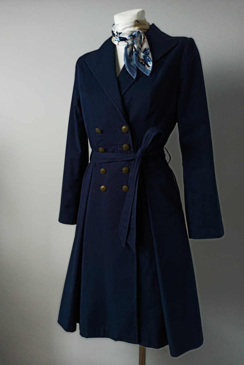 May include: A navy blue double-breasted coat with a belt and gold buttons. The coat has a flared skirt and long sleeves.