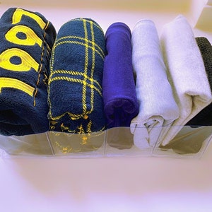 Peut inclure: Une boîte de rangement en plastique transparent contenant cinq vêtements roulés. Les vêtements comprennent une couverture en polaire bleue avec le numéro "1991" brodé dessus, une couverture en plaid bleu et jaune, une couverture en polaire violette, une couverture en polaire grise et une couverture en polaire noire.
