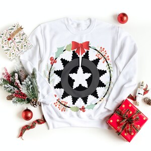 Può includere: Felpa bianca con un disegno di fiocco di neve bianco e nero in una ghirlanda rossa e verde con un fiocco rosso. La felpa è circondata da decorazioni natalizie, tra cui un ornamento rosso, un regalo avvolto e un piccolo albero di pino.