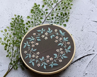 May include: A hand-stitched embroidery hoop featuring a floral wreath design. The wreath is composed of light blue leaves and small white flowers, set against a dark gray fabric. The wooden hoop is visible, and green foliage is in the background.