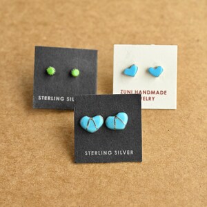 May include: Three pairs of earrings on display. One pair features small, round, green studs. Another pair showcases heart-shaped, turquoise earrings. The third pair has turquoise heart-shaped earrings with silver accents. The cards read "STERLING SILVER" and "ZUNI HANDMADE JEWELRY."