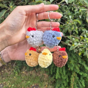 May include: A collection of five small, round, crocheted chicken keychains made from soft, plush yarn. Each chicken features a tiny red comb, a small orange beak, and black bead eyes. The chickens come in grey, blue, tan, yellow, and brown colours.