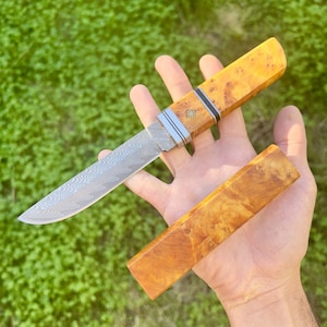 Könnte beinhalten: Ein handgefertigtes Messer mit einer gemusterten Stahlklinge und einem hellen Holzgriff. Der Griff hat ein natürliches Holzmaserungsmuster. Eine passende Holzscheide ist ebenfalls vorhanden. Das Messer wird in einer Hand vor grünem Hintergrund gehalten.
