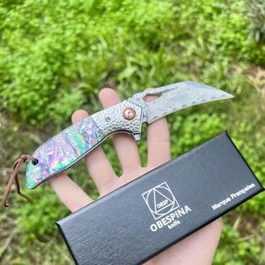 May include: A folding knife with a patterned blade and a handle with iridescent abalone shell inlays. The knife has an orange accent and a leather lanyard. The black box reads "OBESPINA knife" and "Marque Française".