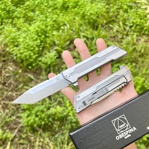 Może przedstawiać: Srebrny nóż kieszonkowy z teksturą na uchwycie i czarne pudełko z napisem "OBESPINA knife" i trójkątnym logo.