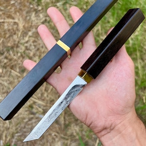 Könnte beinhalten: Ein handgefertigtes Messer mit gemusterter Klinge und dunklem Holzgriff. Das Messer wird mit einer passenden Scheide geliefert, beide mit Messingakzenten. Die Klinge ist etwa 15 cm lang.