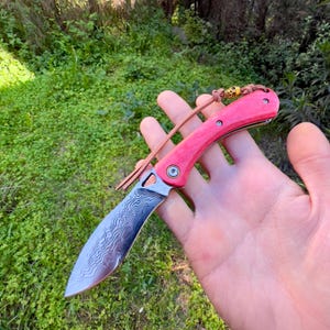 May include: A red and silver pocket knife with a patterned blade and a leather lanyard. The knife is closed and has a skull-shaped bead on the lanyard.