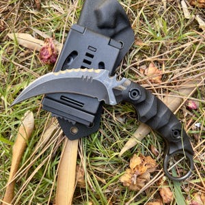 Peut inclure: Un couteau karambit noir et argent avec une lame dentelée et une poignée noire. Le couteau est dans un fourreau en cuir noir avec une boucle de ceinture.