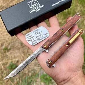 Peut inclure: Un couteau pliant avec une lame en acier Damas et un manche en bois de poirier. Le couteau est ouvert, révélant le motif complexe de la lame. Le manche est de couleur marron foncé et le couteau est tenu dans une main. Le couteau est de la marque Obespina.