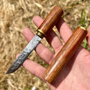 Peut inclure: Un petit couteau décoratif avec une lame en acier à motifs et un manche en bois. Le manche est cylindrique et possède un raccord en laiton. Le couteau est présenté avec son étui en bois, également cylindrique.