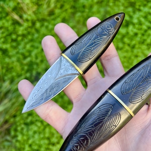 May include: A close-up of two small, decorative knives. The blades have a patterned design, and the handles are dark with a cicada engraving. A gold-colored band accents the blade and handle connection. The knives are held against a blurred green background.
