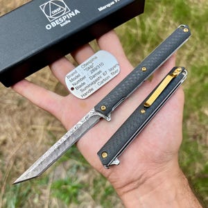 Può includere: Un coltello OBESPINA nero con manico in fibra di carbonio e lama Damasco. Il coltello è aperto, rivelando l'intricato motivo della lama. Il testo sull'etichetta recita "Brand: Obespina, Model: Skip, Blade: Damas".