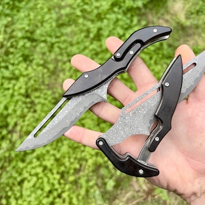 May include: Two folding pocket knives with black wood handles and patterned steel blades. The blades are partially open and the knives are held in a hand.