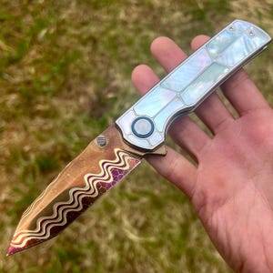 May include: A folding pocket knife with a metallic blade featuring a wavy, multi-colored pattern of gold, bronze, and purple with a hexagonal texture. The handle is constructed from silver-toned metal segments inlaid with iridescent, pearlescent panels.