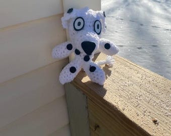 May include: A white crocheted dalmatian dog plush toy with black spots and large black eyes. The toy is sitting on a wooden railing.
