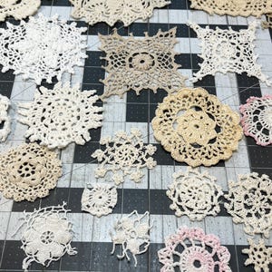 May include: A collection of various handmade crochet doilies in shades of white, cream, and light pink. The doilies feature intricate floral and geometric patterns, showcasing detailed craftsmanship. Some are circular, others square, and some have scalloped edges.