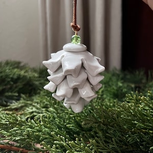 May include: A white ceramic ornament shaped like a pinecone, suspended by a brown cord. A small green bead accents the top. The ornament is displayed against a backdrop of green foliage, suggesting a holiday theme.