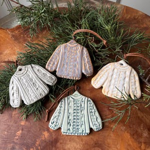 May include: Four ceramic sweater ornaments in cream, light blue, and grey, with brown leather hanging loops. The ornaments are displayed on a wooden surface with green sprigs. Each sweater has a unique textured design.