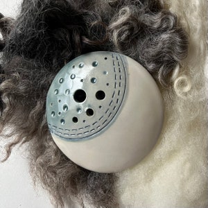 May include: A ceramic, round object with a light blue, perforated top and a cream-coloured base. The top has a dotted pattern and a stitched design. The object is set against a backdrop of grey and white wool.