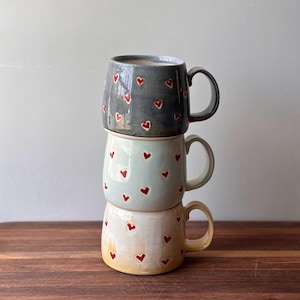 May include: Stack of three ceramic mugs with red heart patterns. The top mug is gray, the middle mug is light blue, and the bottom mug is cream.
