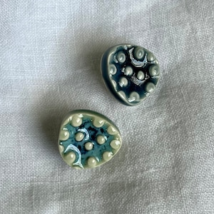 May include: Two ceramic buttons with a blue and white speckled design. The buttons are shaped like a rounded oval with a raised, textured surface.