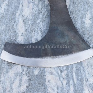 May include: Close-up of a black axe head with a slightly curved blade and a polished edge. The axe head is resting on a light gray marble surface. The text "antiquegiftcraftsCo" is visible on the axe head.