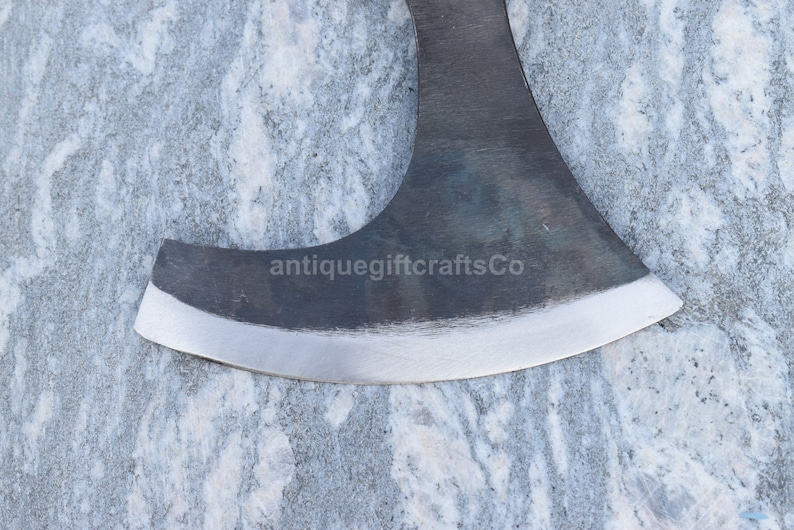 May include: Close-up of a black axe head with a slightly curved blade and a polished edge. The axe head is resting on a light gray marble surface. The text "antiquegiftcraftsCo" is visible on the axe head.