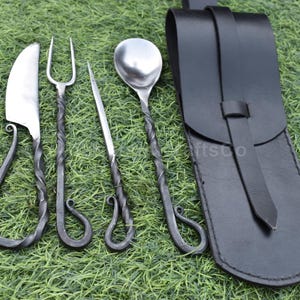 May include: A medieval-style cutlery set with a knife, fork, spoon, and skewer, all with twisted handles and a black leather sheath. The utensils are made of silver-colored metal.