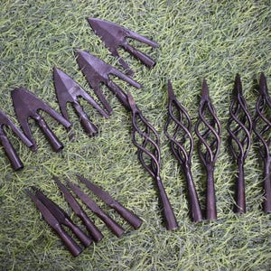 May include: A collection of 10 arrowheads, including 6 with a traditional design and 4 with a twisted, cage-like design. The arrowheads are made of metal and have a dark brown finish.