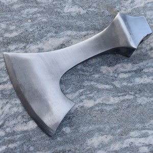 May include: A silver axe with a wide, curved blade and a straight handle. The axe is lying on a gray and white surface.