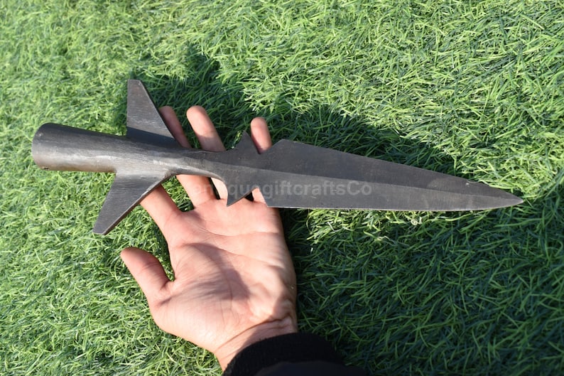K&ouml;nnte beinhalten: Ein dunkelgrauer Speerspitze mit einer langen, spitzen Klinge und einem markanten fl&uuml;gelf&ouml;rmigen Design nahe der Basis. Der Speerspitze wird vor einem Hintergrund aus gr&uuml;nem Gras gehalten. Der Text "quagiftcraftsCo" ist sichtbar.