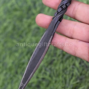 Può includere: Un pugnale in metallo nero con un'impugnatura attorcigliata e una lama affilata e appuntita. Il testo "antiquegiftcraftsCo" è visibile sulla lama. Il pugnale è tenuto su uno sfondo verde sfocato.