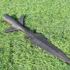 K&ouml;nnte beinhalten: Ein schwarzer Speerkopf mit einem markanten Design, der eine zentrale Klinge und fl&uuml;gelf&ouml;rmige Vorspr&uuml;nge aufweist. Der Speerkopf besteht aus dunklem Metall und liegt auf gr&uuml;nem Gras.