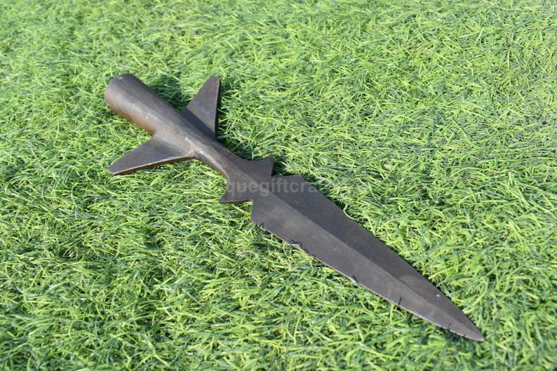 K&ouml;nnte beinhalten: Ein schwarzer Speerkopf mit einem markanten Design, der eine zentrale Klinge und fl&uuml;gelf&ouml;rmige Vorspr&uuml;nge aufweist. Der Speerkopf besteht aus dunklem Metall und liegt auf gr&uuml;nem Gras.