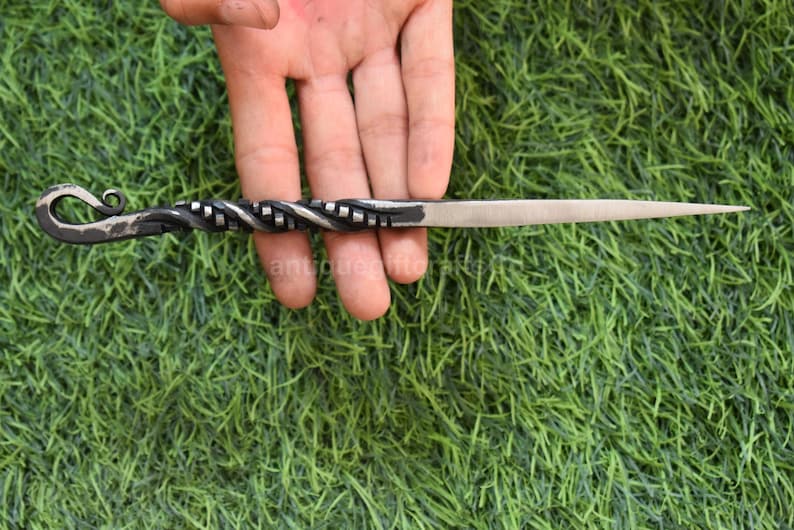 May include: A hand-forged letter opener with a twisted handle and a pointed blade. The handle features a decorative spiral design and a curled end. The metal has a dark, textured finish, and the blade is silver.