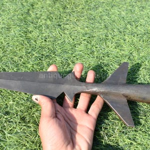 K&ouml;nnte beinhalten: Ein dunkler Metall-Speerkopf mit einer scharfen, spitzen Klinge und einem zylindrischen Griff. Der Speerkopf hat zwei Flossen und eine gezackte Kante. Das Objekt wird vor einem Hintergrund aus gr&uuml;nem Gras gehalten.