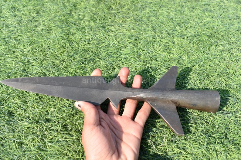 K&ouml;nnte beinhalten: Ein dunkler Metall-Speerkopf mit einer scharfen, spitzen Klinge und einem zylindrischen Griff. Der Speerkopf hat zwei Flossen und eine gezackte Kante. Das Objekt wird vor einem Hintergrund aus gr&uuml;nem Gras gehalten.