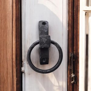 Puede incluir: Un aldaba de puerta de hierro forjado negro con un asa circular y una placa de montaje rectangular. La aldaba está montada en una puerta blanca con un marco de madera. El texto "antiquegiftcrafts Co" es visible.
