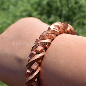 Puede incluir: Una pulsera de color cobre con un diseño geométrico. La pulsera se lleva en el brazo de una persona. El texto "antiquecraftsCo" es visible en el brazo. El fondo es hierba verde borrosa.