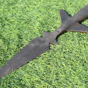 K&ouml;nnte beinhalten: Ein dunkelbrauner Speerkopf mit einer langen, spitzen Klinge und einem markanten, stilisierten Design. Der Speerkopf liegt auf einem Bett aus gr&uuml;nem Gras. Der Speerkopf hat eine einzigartige Form mit einer zentralen Klinge und fl&uuml;gelf&ouml;rmigen Vorspr&uuml;ngen.
