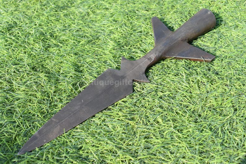 K&ouml;nnte beinhalten: Ein dunkelbrauner Speerkopf mit einer langen, spitzen Klinge und einem markanten, stilisierten Design. Der Speerkopf liegt auf einem Bett aus gr&uuml;nem Gras. Der Speerkopf hat eine einzigartige Form mit einer zentralen Klinge und fl&uuml;gelf&ouml;rmigen Vorspr&uuml;ngen.