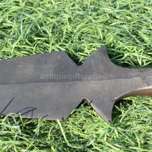 K&ouml;nnte beinhalten: Nahaufnahme einer dunklen, klingenartigen Waffe mit gezackter Kante, m&ouml;glicherweise ein Schwert oder ein gro&szlig;es Messer. Die Waffe liegt auf einem Bett aus gr&uuml;nem Gras. Der Text "antiquegiftcraftsCo" ist auf der Klinge zu sehen.