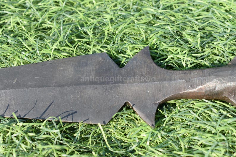 K&ouml;nnte beinhalten: Nahaufnahme einer dunklen, klingenartigen Waffe mit gezackter Kante, m&ouml;glicherweise ein Schwert oder ein gro&szlig;es Messer. Die Waffe liegt auf einem Bett aus gr&uuml;nem Gras. Der Text "antiquegiftcraftsCo" ist auf der Klinge zu sehen.