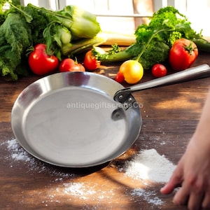 Könnte beinhalten: Eine Edelstahl-Bratpfanne mit langem Griff, die auf einer Holzoberfläche liegt. Frisches Gemüse, darunter Tomaten und Blattgemüse, ist um die Pfanne herum angeordnet. Das Bild enthält den Text "antiquegiftscrapsCo".